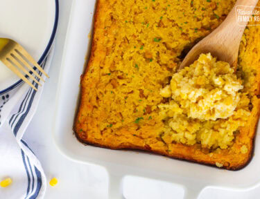 Wooden spoon with scooped corn casserole in a baking dish.