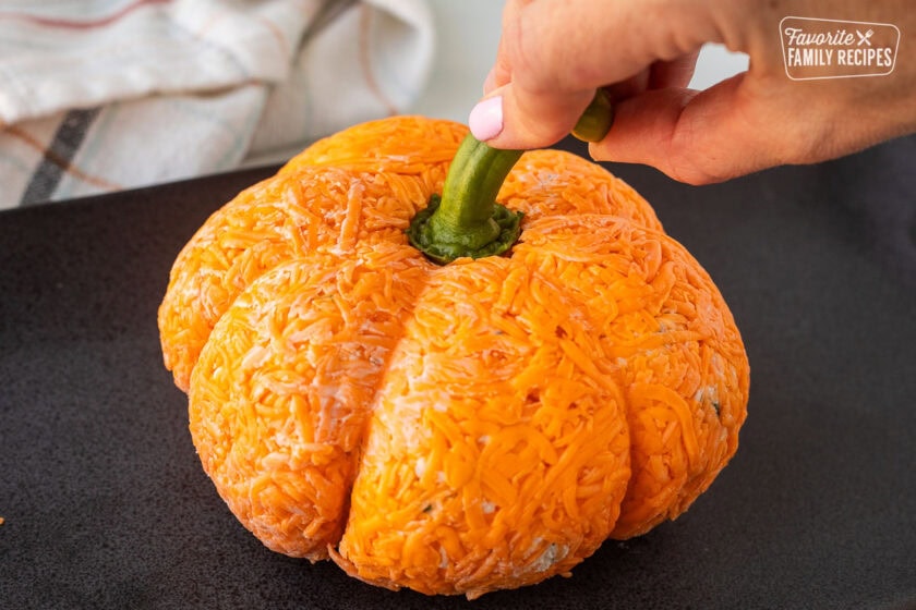 Hand pushing a bell pepper stem on top of a pumpkin shaped cheese ball.