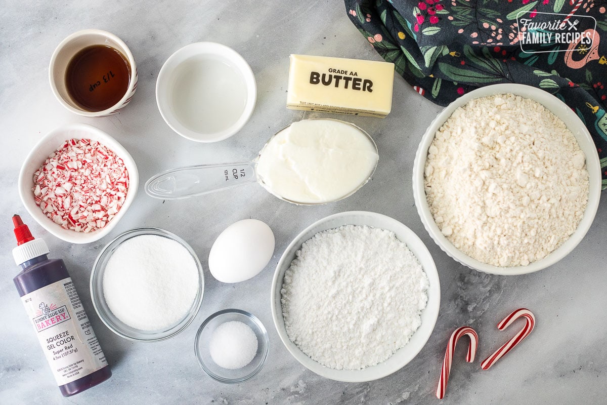 Ingredients to make candy, cane, cookies, including flour, powdered sugar, sugar, salt, egg, shortening, butter, almond extract, vanilla, crushed candy, canes, and red gel color.