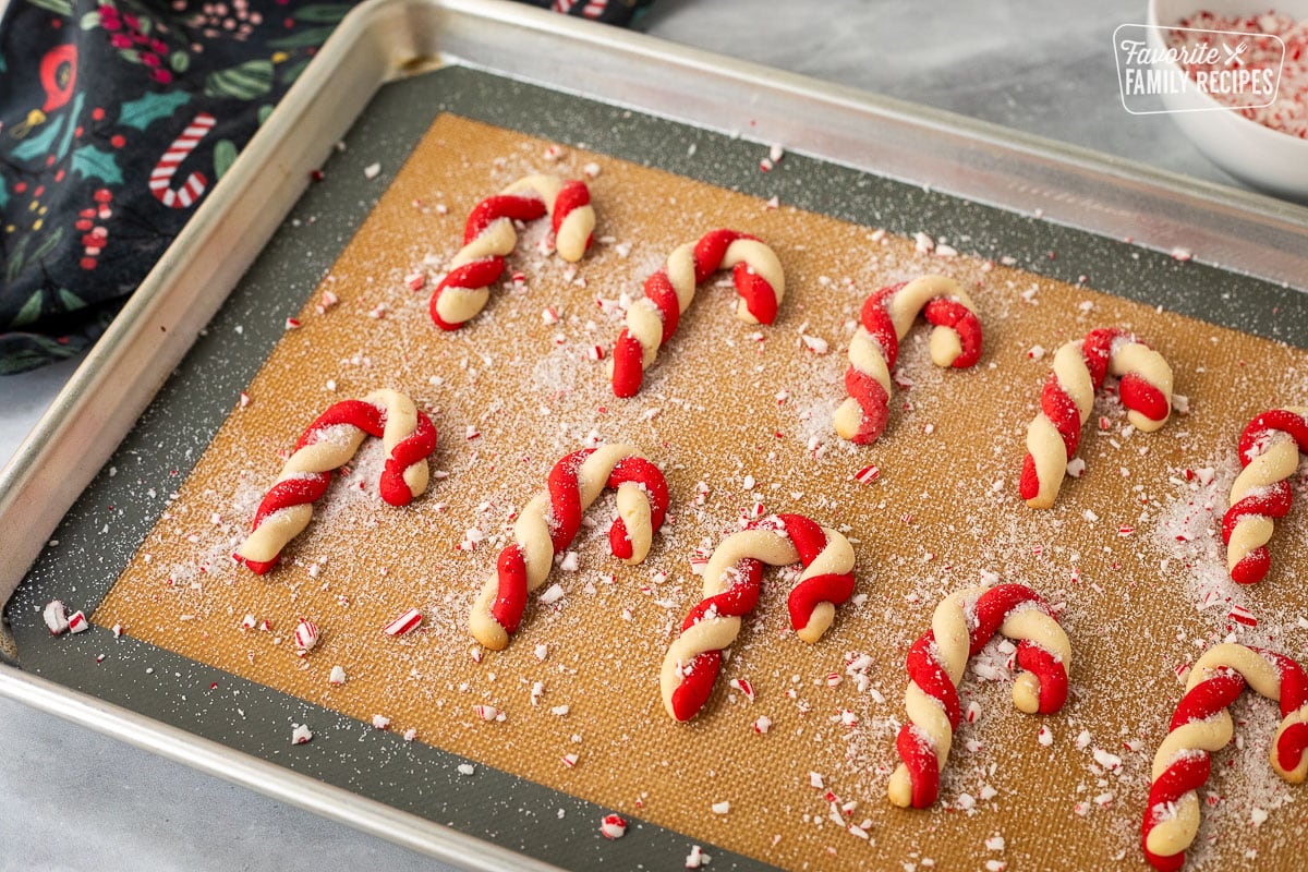 Candy cane cookies sprinkled with crushed peppermint, candies, and sugar on a cookie sheet.