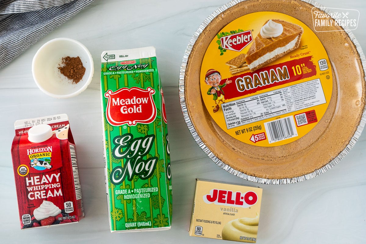 Ingredients to make egg nog pie, including graham cracker crust, eggnog, vanilla, pudding, pumpkin pie spice, and heavy whipping cream.