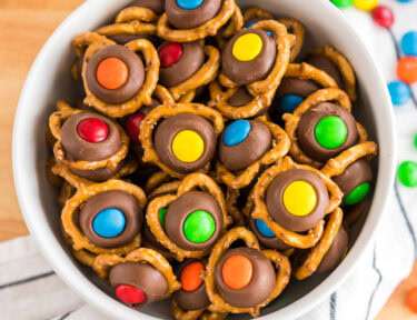 Hershey Kiss Pretzels in a white bowl on a white linen cloth with gray stripes. M&M's are sprinkled on the linen.