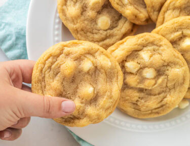 Soft and buttery white chocolate chip cookies on a platter with one cookie being taken from the plate.