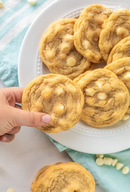 Soft and buttery white chocolate chip cookies on a platter with one cookie being taken from the plate.