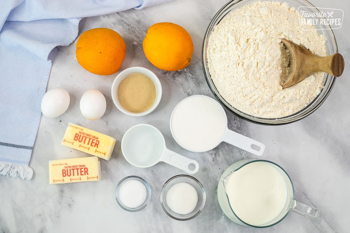 Ingredients to make homemade orange rolls, including flour, oranges, yeast, sugar, milk, water, salt, eggs, and butter.