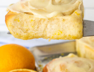 Frosted homemade orange roll on a spatula over a pan of orange rolls.