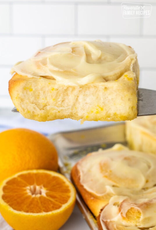 Frosted homemade orange roll on a spatula over a pan of orange rolls.