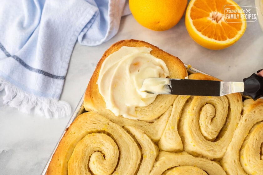 Spreading frosting onto baked orange rolls with a spatula.