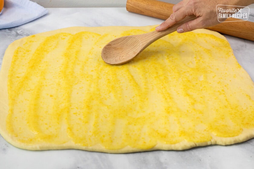 Spreading orange glaze on rolled out bread dough with a wooden spoon.