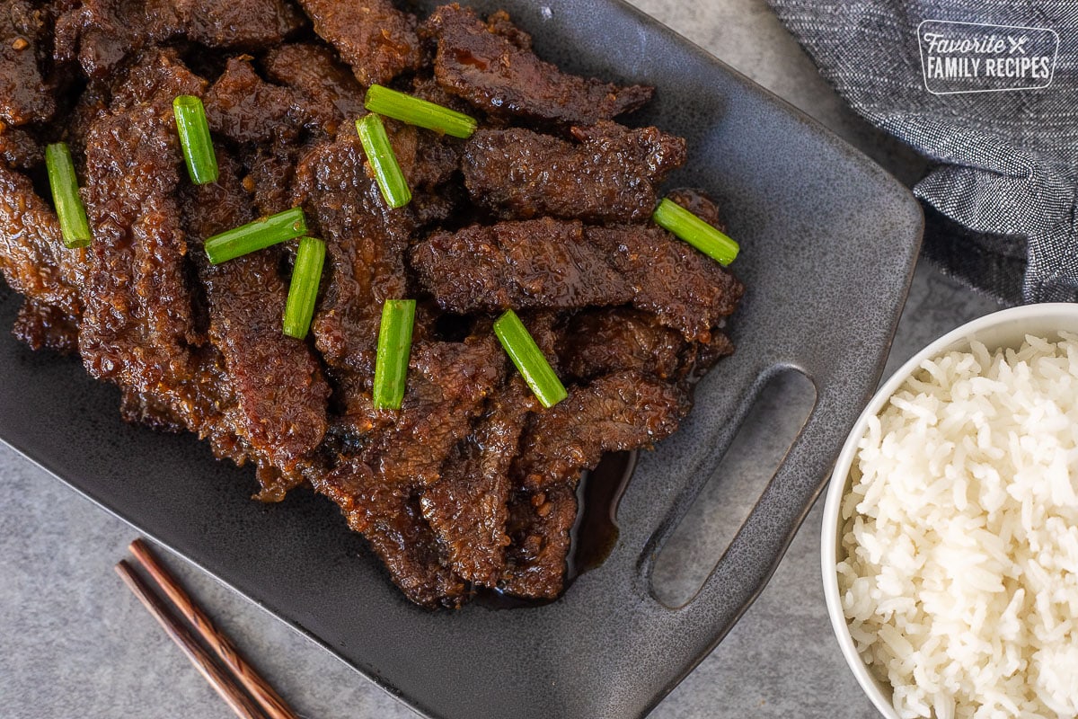 Mongolian beef on a platter with green onion and a bowl of rice on the side.