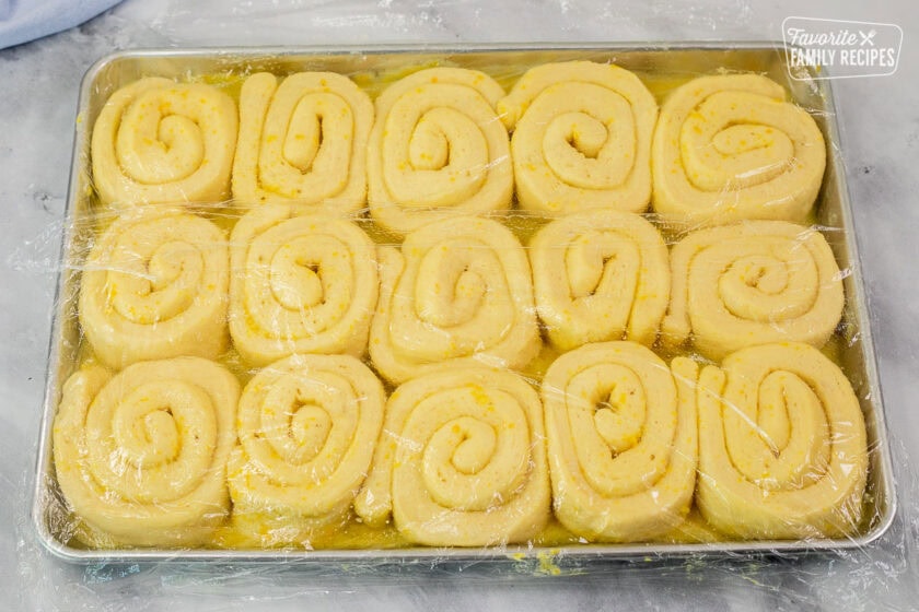 Homemade orange rolls rising in baking sheet covered in plastic wrap.