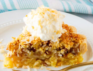 Slice of Island Pecan Pie on a plate topped with a coconut cream and toasted coconut. Fork resting on the plate.