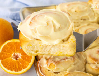 Frosted homemade orange roll on a spatula over a pan of orange rolls.