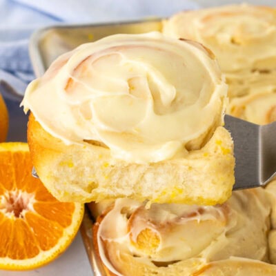 Frosted homemade orange roll on a spatula over a pan of orange rolls.