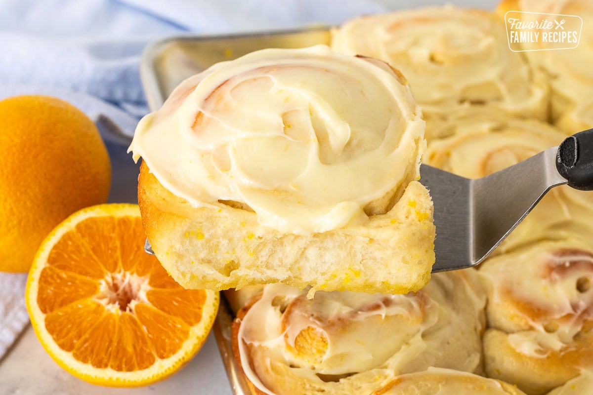 Frosted homemade orange roll on a spatula over a pan of orange rolls.