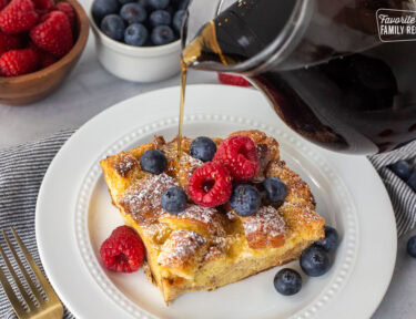 Pouring syrup on top of a slice of brioche French toast casserole topped with fresh blueberries and raspberries.