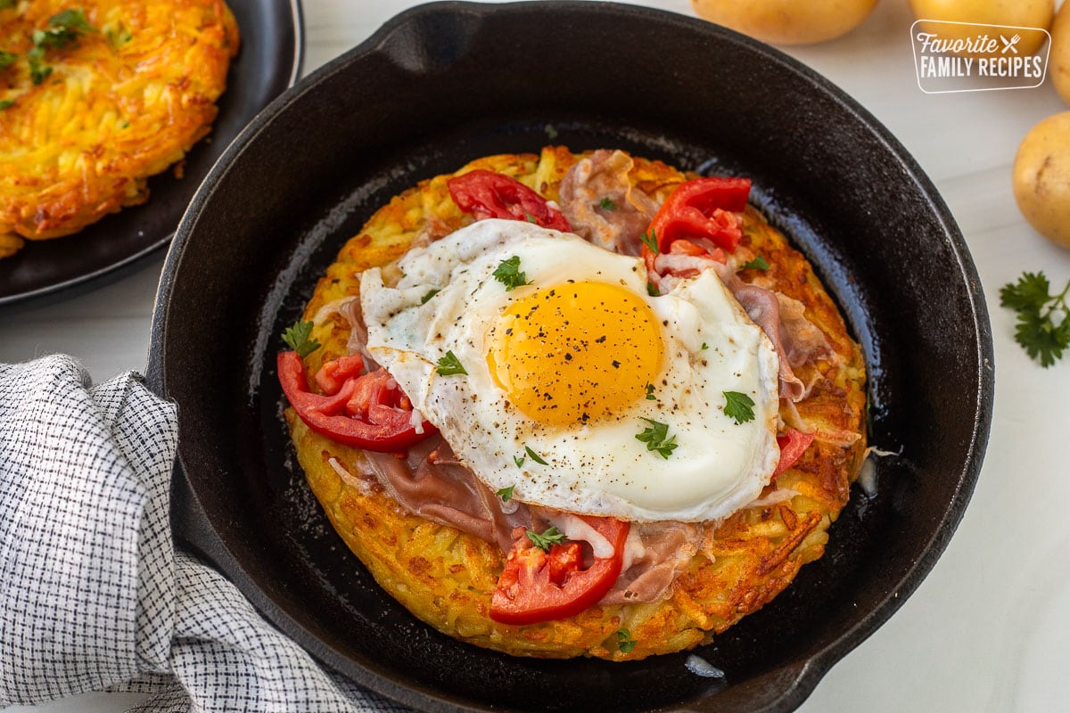 Rosti in a skillet topped with cheese, prosciutto, tomatoes, egg and parsley and rosti on a plate.