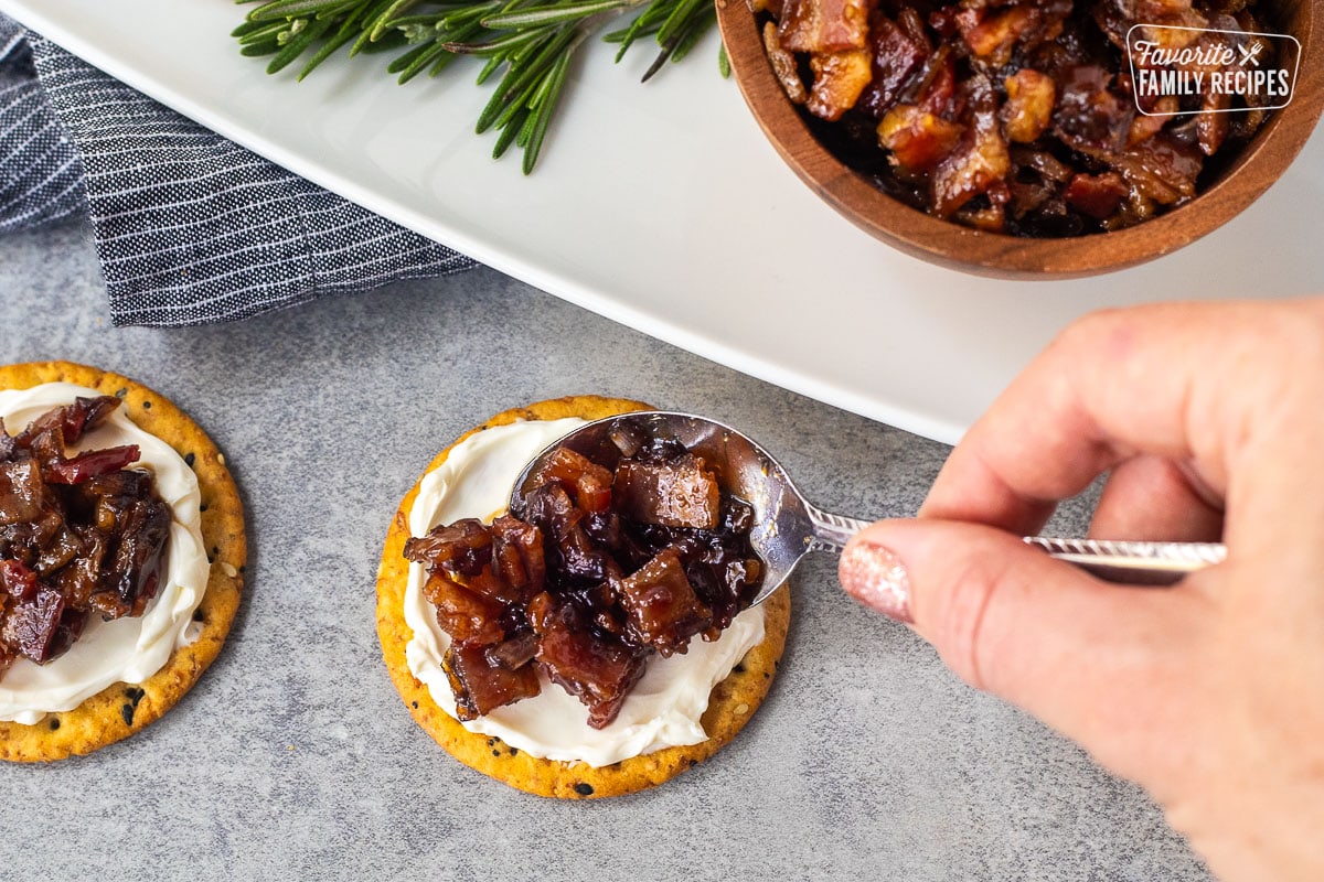 Pouring bacon jam over a cracker spread with cream cheese.