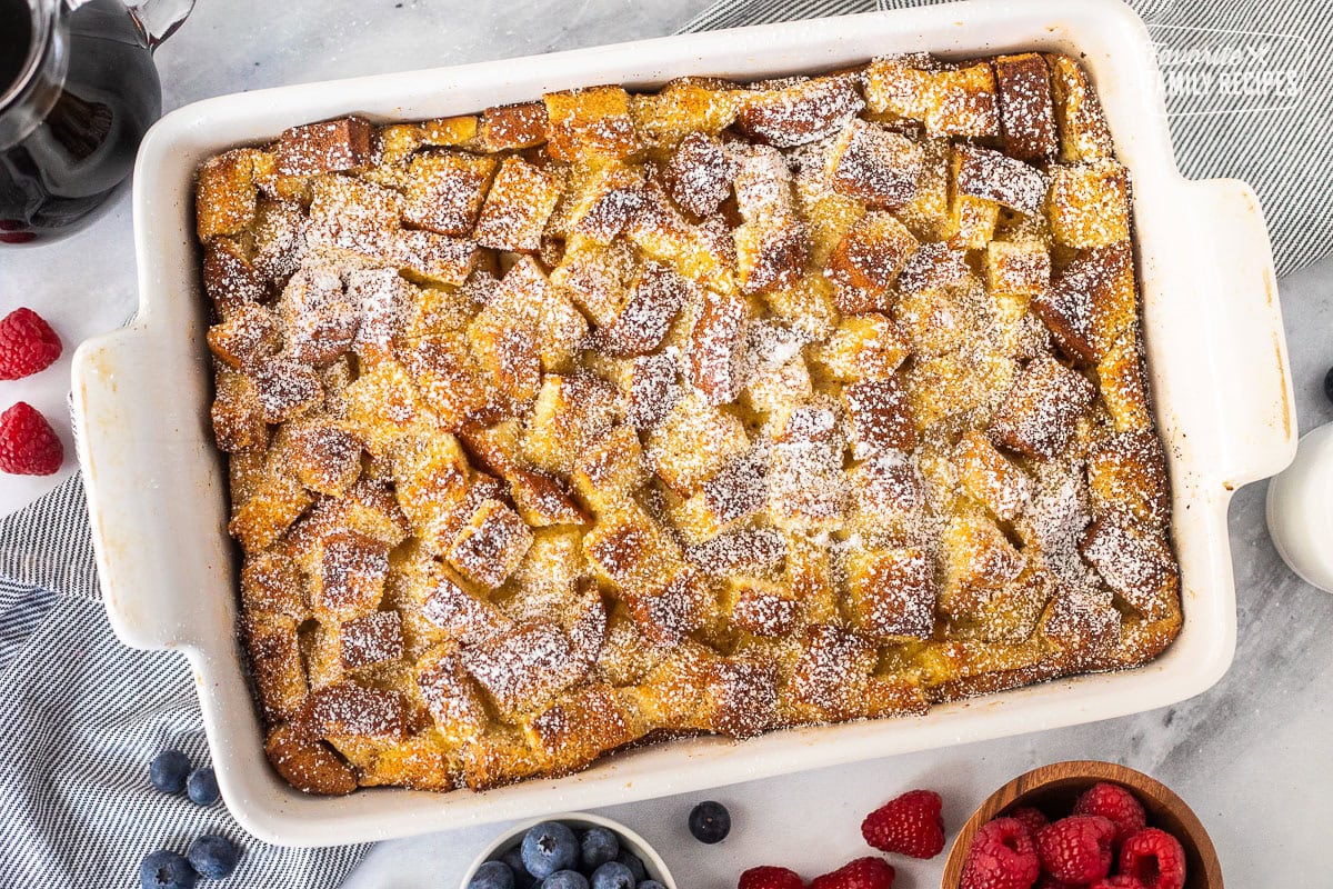 Baking dish of brioche French toast casserole dusted with powdered sugar. Blueberries and raspberries on the side.