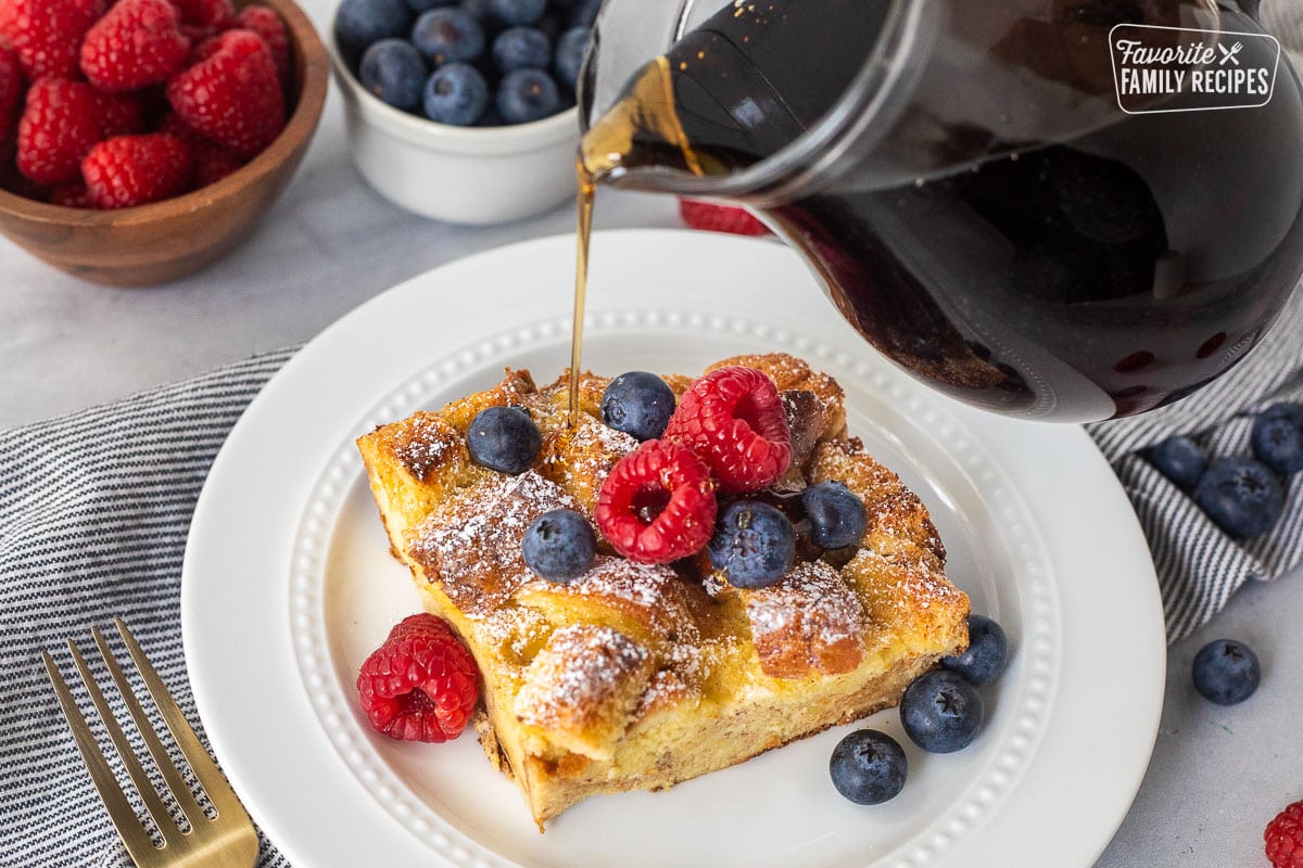 Pouring syrup on top of a slice of brioche French toast casserole topped with fresh blueberries and raspberries.