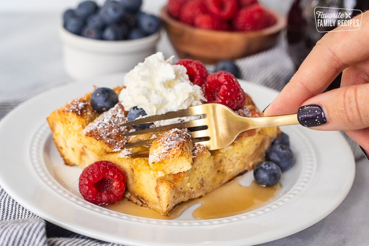 Fork cutting into a corner of brioche French toast casserole slice topped with berries and whipped cream.