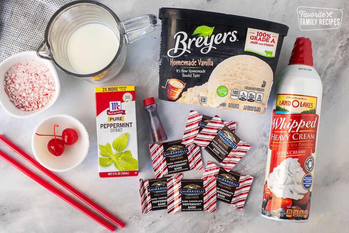 Ingredients to make Chick Fil A Peppermint Milkshakes including vanilla ice cream, Ghirardelli dark chocolate peppermint bark, peppermint extract, crushed candy, canes, milk, whipped cream, cherries, red food coloring, and straws.