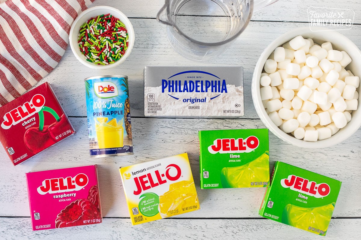 Ingredients to make Christmas Jell-O salad, including cream cheese, mini marshmallows, water, sprinkles, pineapple juice, lime Jell-O, lemon Jell-O, raspberry Jell-O, and cherry Jell-O.