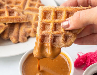 Holding a churro waffle piece dipped in caramel sauce. Plate of churro waffles and fresh strawberries in the background.