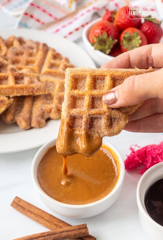 Holding a churro waffle piece dipped in caramel sauce. Plate of churro waffles and fresh strawberries in the background.