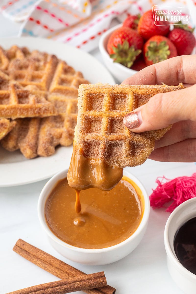 Holding a churro waffle piece dipped in caramel sauce. Plate of churro waffles and fresh strawberries in the background.