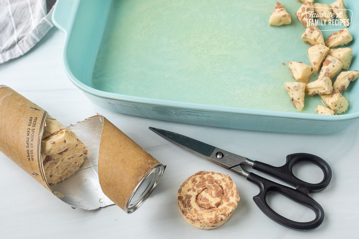 Cinnamon rolls in a can next to a pair of kitchen shears. Baking dish with butter and cut up pieces of cinnamon roll.