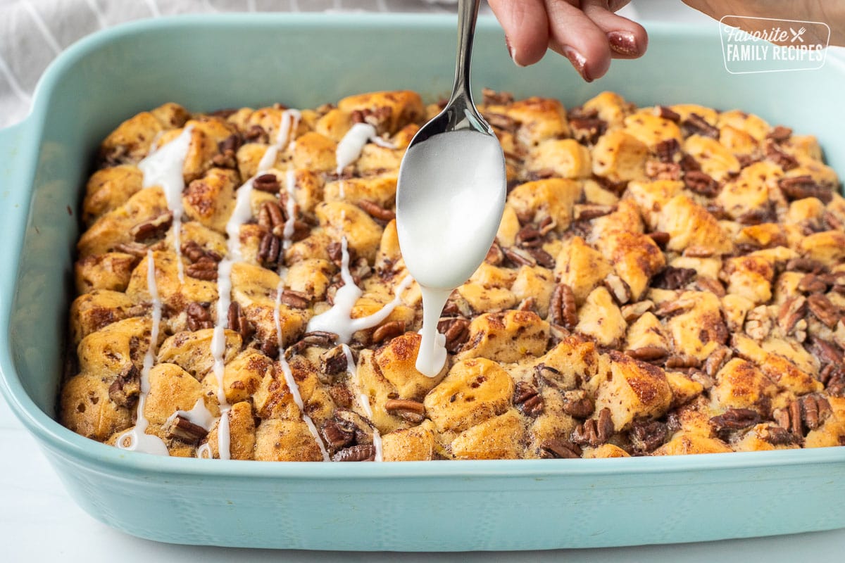 Cinnamon Roll Casserole in a baking dish with pecans on top drizzling white icing on top with a spoon.