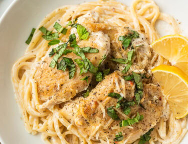 Plate with crockpot garlic Parmesan chicken over pasta with fresh lemon wedges and basil garnished on top.