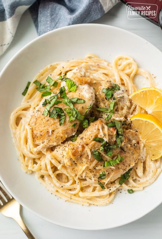 Plate with crockpot garlic Parmesan chicken over pasta with fresh lemon wedges and basil garnished on top.