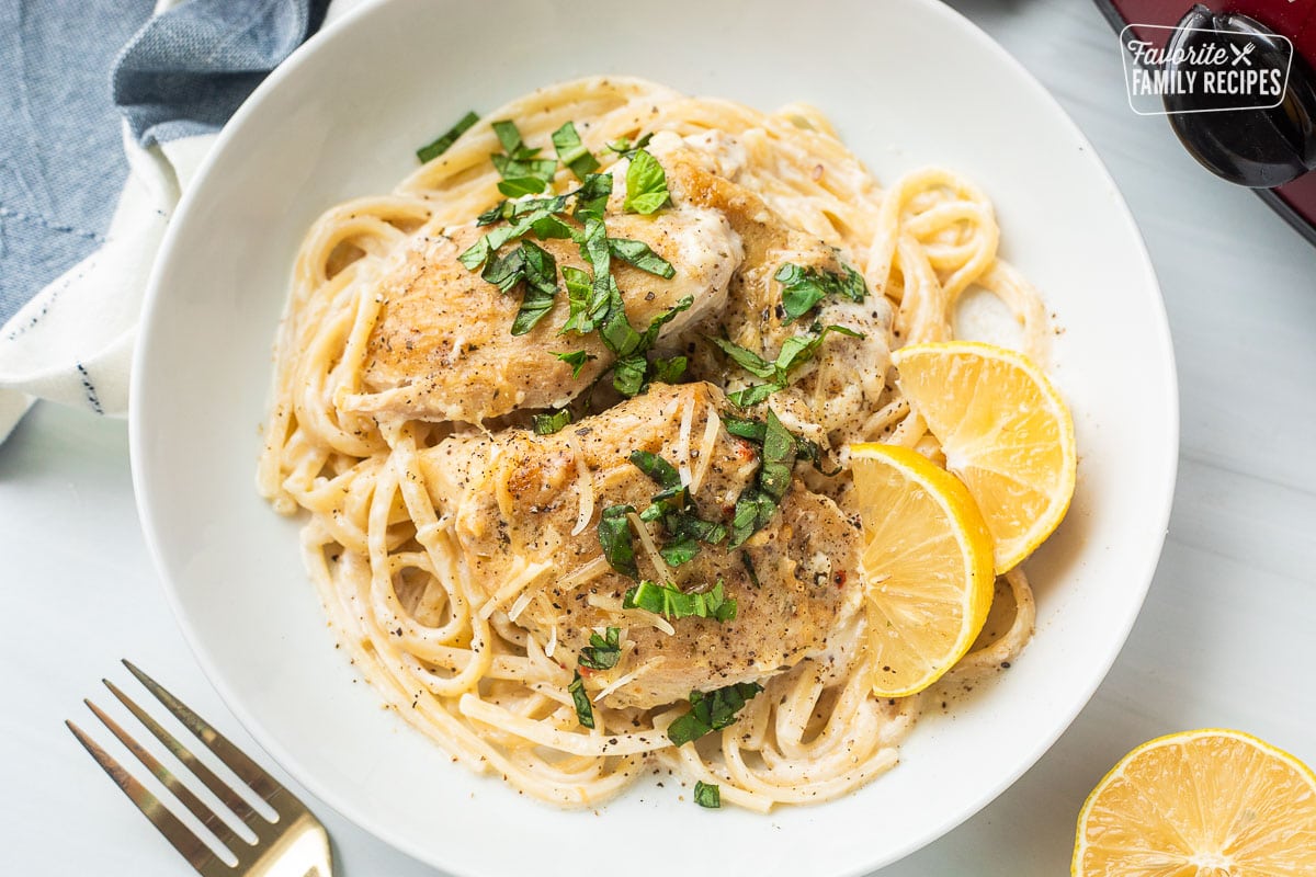 Plate with crockpot garlic Parmesan chicken over pasta with fresh lemon wedges and basil garnished on top. Crockpot in the background.