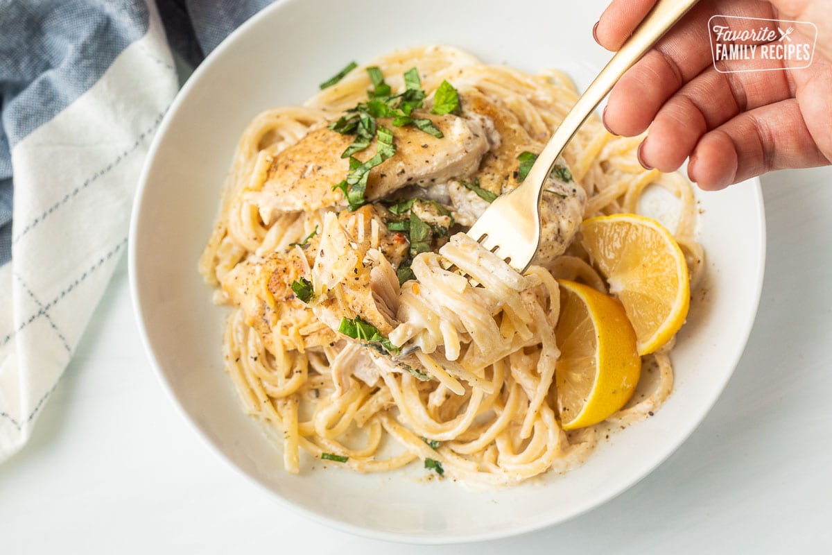 Fork twirling pasta with creamy garlic Parmesan chicken garnished with basil and lemon wedges.