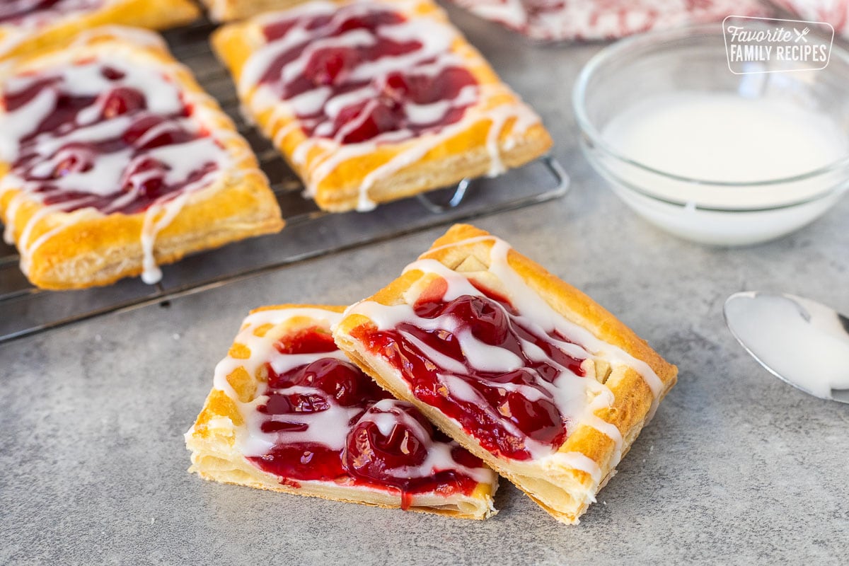 Cherry danish, cut and half with white icing. Cooling rack of cherry danishes in the background with a dish of white icing and spoon.