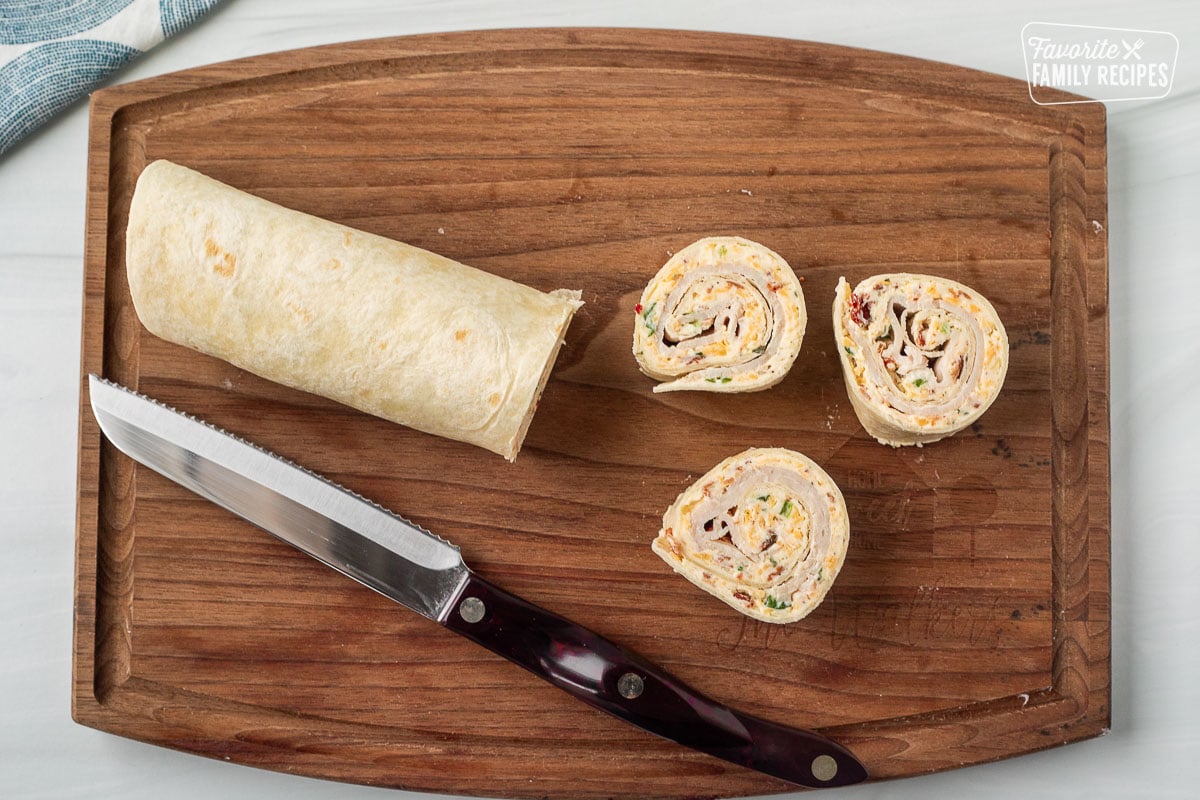 An image of a knife on a cutting board in the process of slicing sections of turkey pinwheels from a filled and rolled tortilla