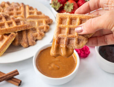 Holding a churro waffle piece dipped in caramel sauce. Plate of churro waffles and fresh strawberries in the background.