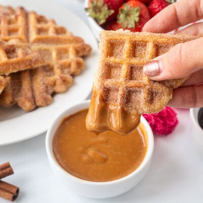 Holding a churro waffle piece dipped in caramel sauce. Plate of churro waffles and fresh strawberries in the background.