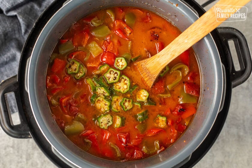 Instant pot with okra added to gumbo ingredients and a wooden spoon.