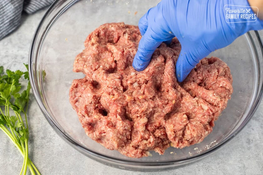 Gloved hand mixing meatball ingredients together in a glass mixing bowl.