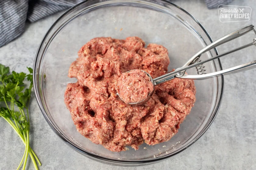 Cookie scoop filled with meatball mixture resting in a glass mixing bowl.