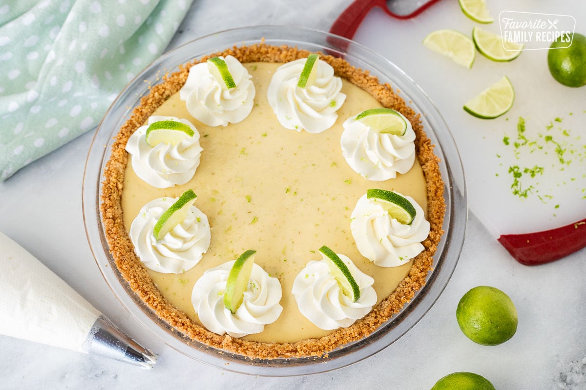 A whole key lime pie decorated with whipped cream and thin slices of lime over the top next to a cutting board of sliced limes