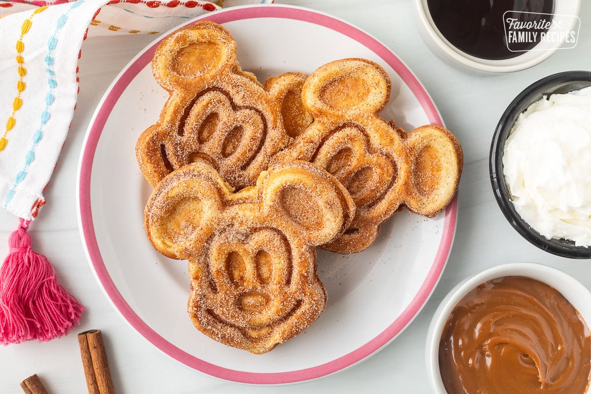 Mickey Mouse shaped churro waffles on a plate. Sides of chocolate dipping sauce, caramel dipping sauce and whipped cream cream.