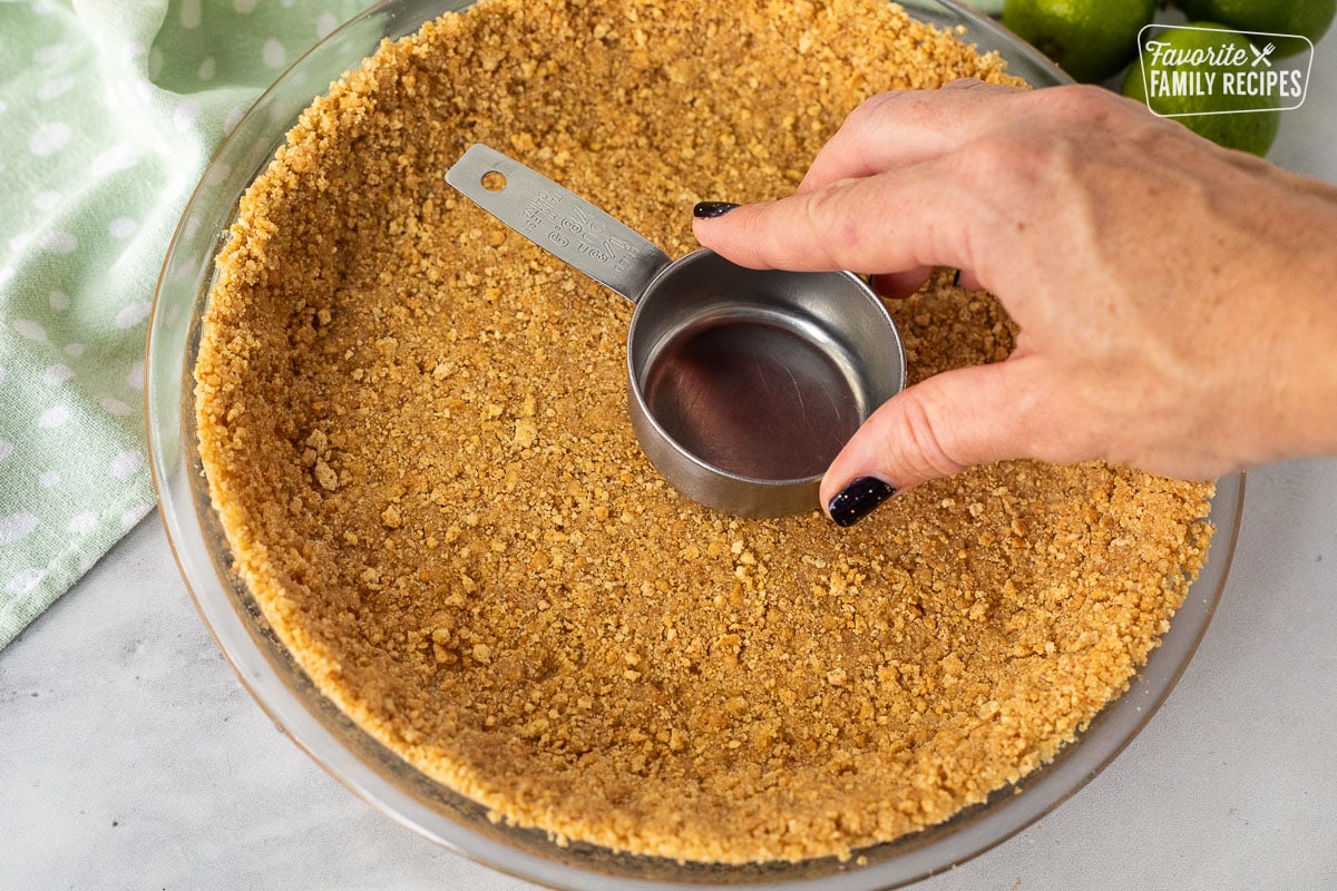 A graham cracker crust being pressed down with the back of a measuring cup, making the bottom of the pie crust flat.