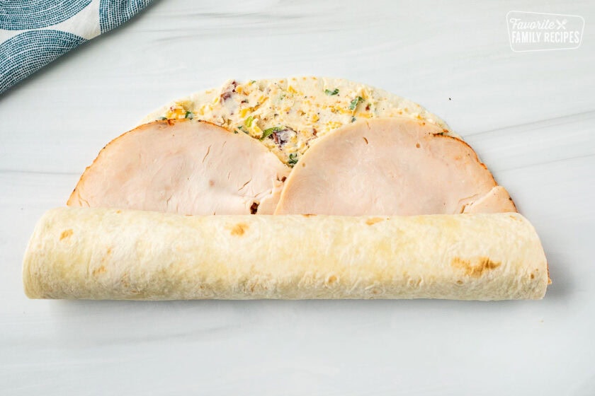 A tortilla with a filling made from cream cheese and turkey slices being rolled up.