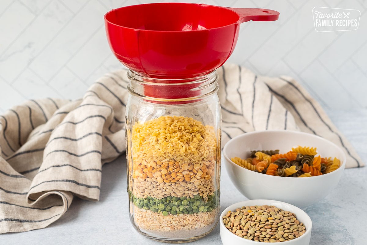 Quart size jar with layered beef bouillon, dried onions, split green peas, barley, yellow peas, alphabet pasta with a funnel on top. Bowls of tri colored pasta and lentils on the side.