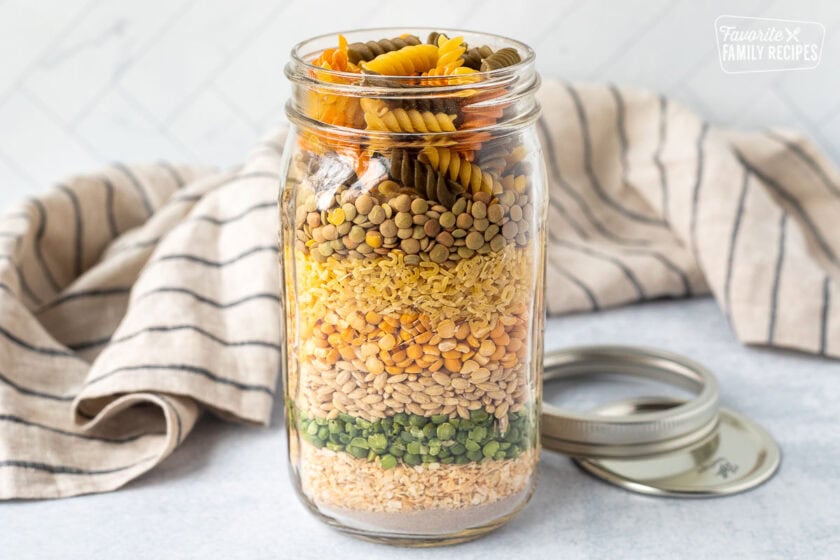 Quart sized jar with layered ingredients to make soup including beef bouillon, dried onions, split green peas, barley, yellow peas, alphabet pasta, lentils and tri colored pasta. Lid and ring on the side.