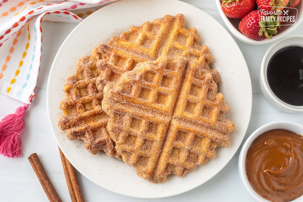 Three churro waffles on a plate next to a small bowl of fresh strawberries, chocolate dipping sauce, and caramel dipping sauce.